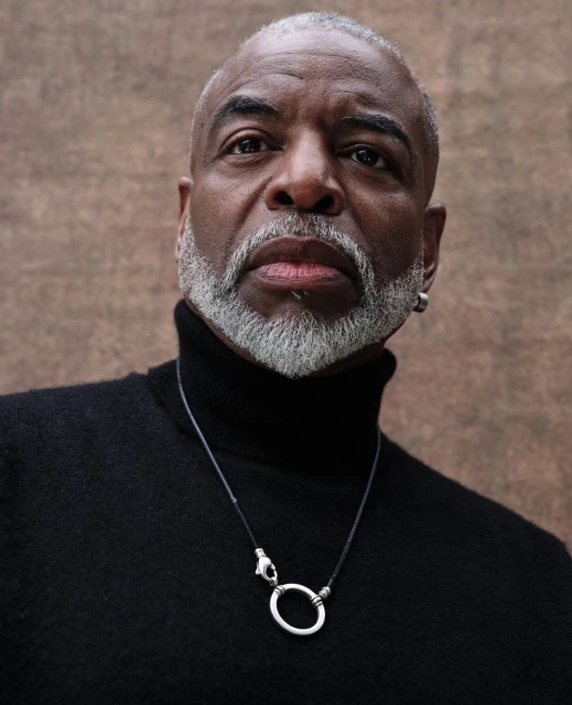 Portrait of a mature adult African American man wearing a black turtleneck and silver necklace against a mottled brown background.