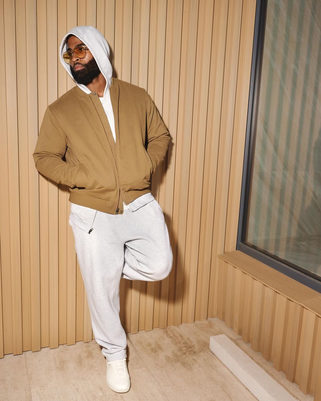 Man wearing a brown bomber jacket, light grey hoodie and sweatpants leaning against a slatted wooden wall by a window.