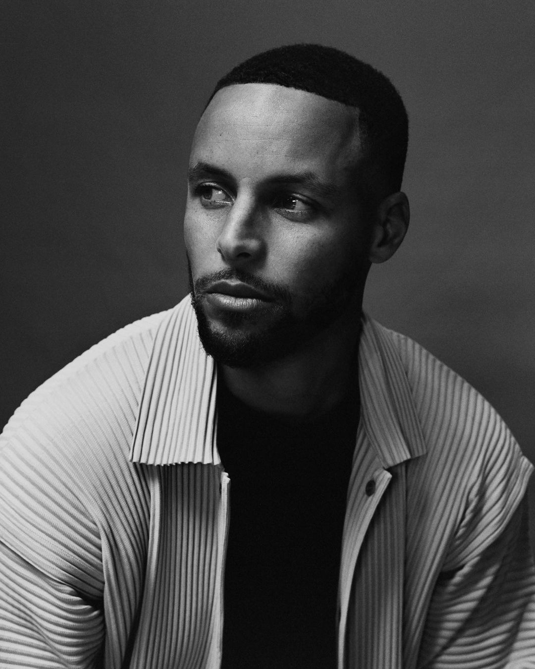 Monochrome portrait of Stephen Curry looking away, wearing a pleated button-up shirt over a dark t-shirt against a plain studio background.