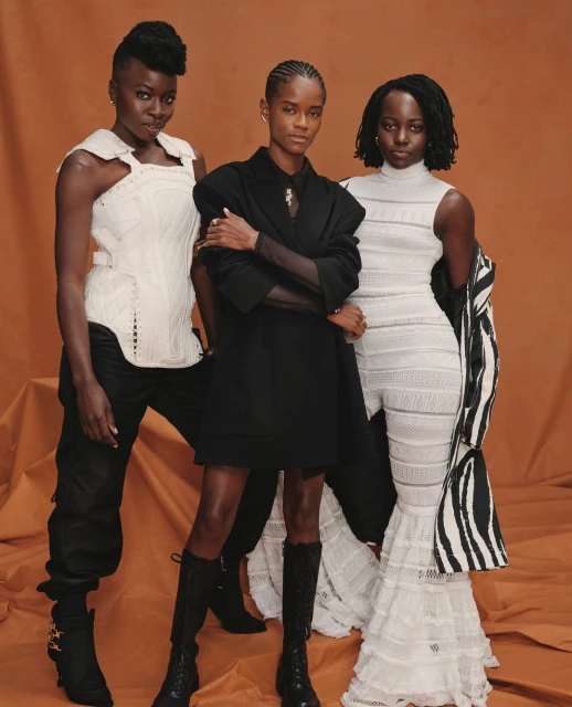 Three African women pose in coordinated studio fashion. One wears white corset and black pants, one a black mini dress, one a white dress.