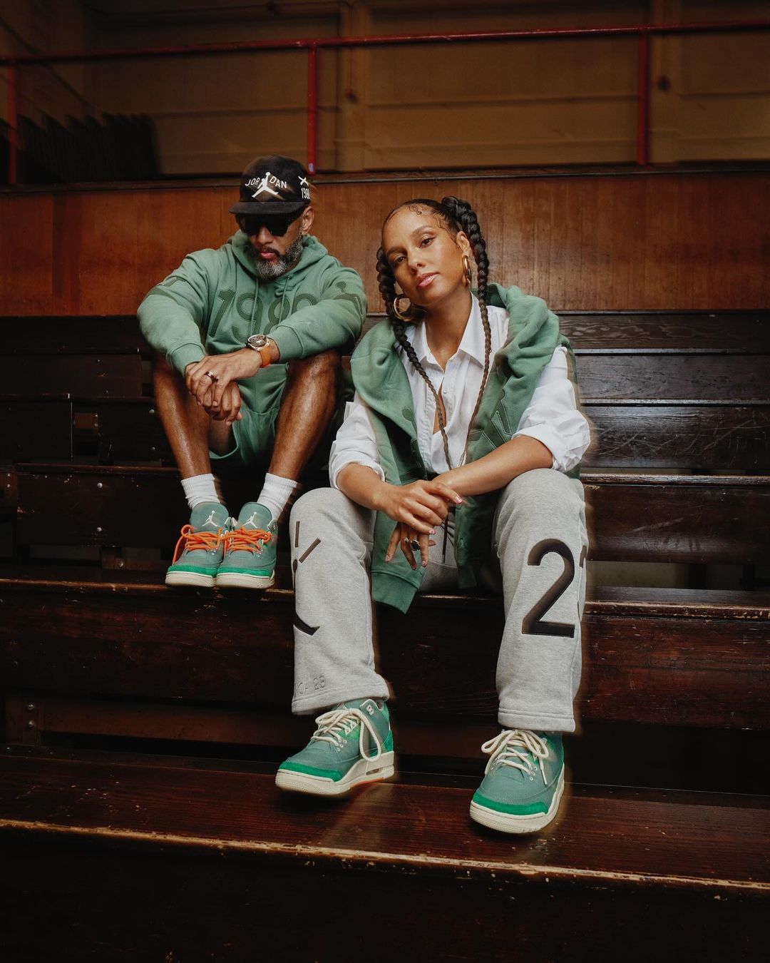 Two people sitting on stadium stairs in green and grey outfits, including hoodies, sweatpants, and sneakers, in an indoor environment.