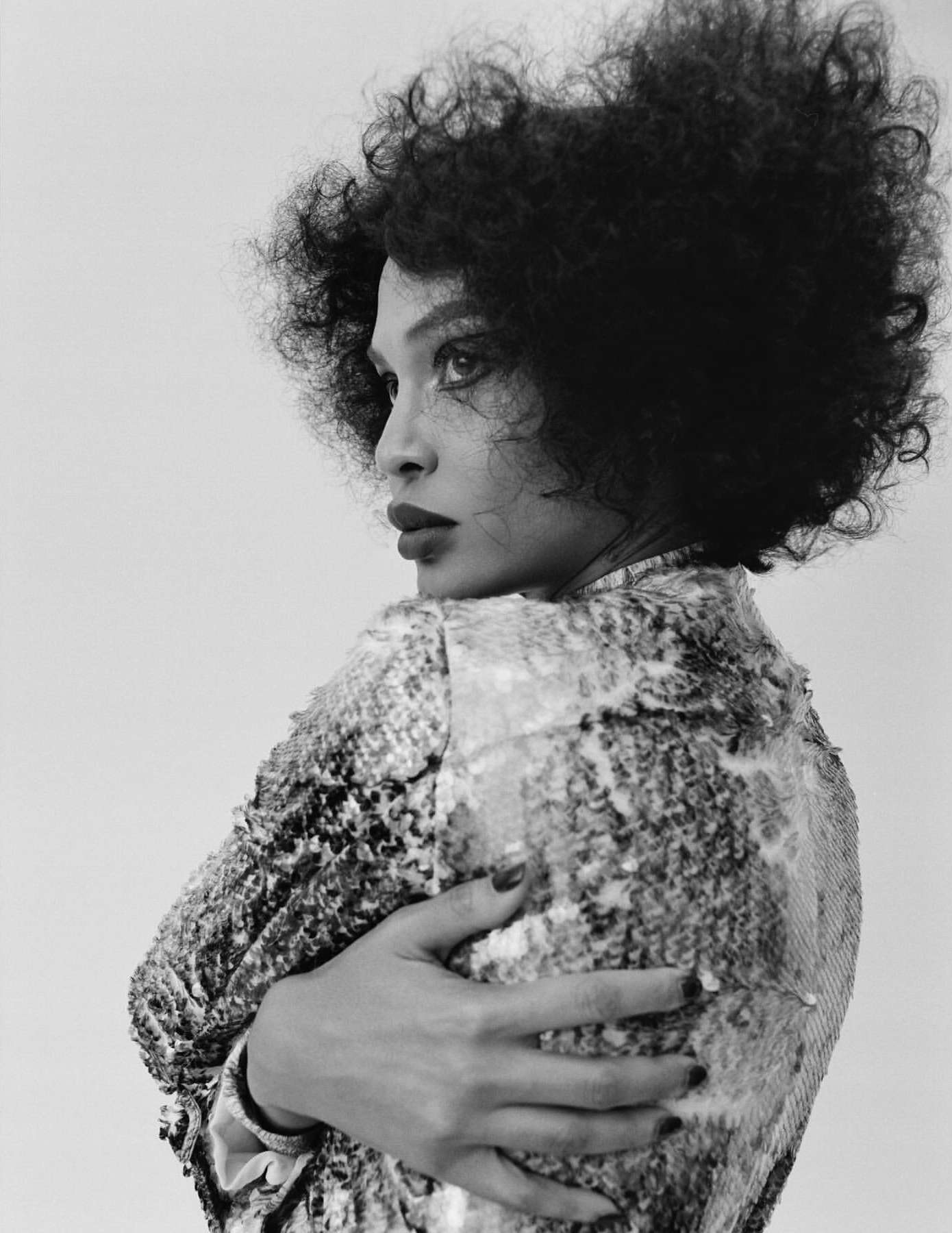 Black and white portrait of a woman with curly hair, wearing a sequined jacket, looking over her shoulder in a studio setting.