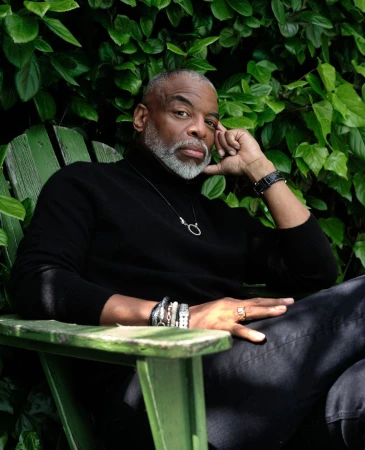 Portrait of an older Black man with a silver beard wearing a black turtleneck seated on a green wooden chair outdoors against a lush hedge