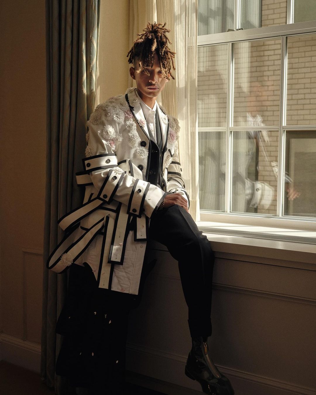 A young adult poses indoors by a window in a stylish, multi-layered fashion jacket with hair in dreadlocks, wearing dress pants and boots.