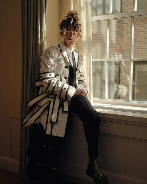A young adult poses indoors by a window in a stylish, multi-layered fashion jacket with hair in dreadlocks, wearing dress pants and boots.