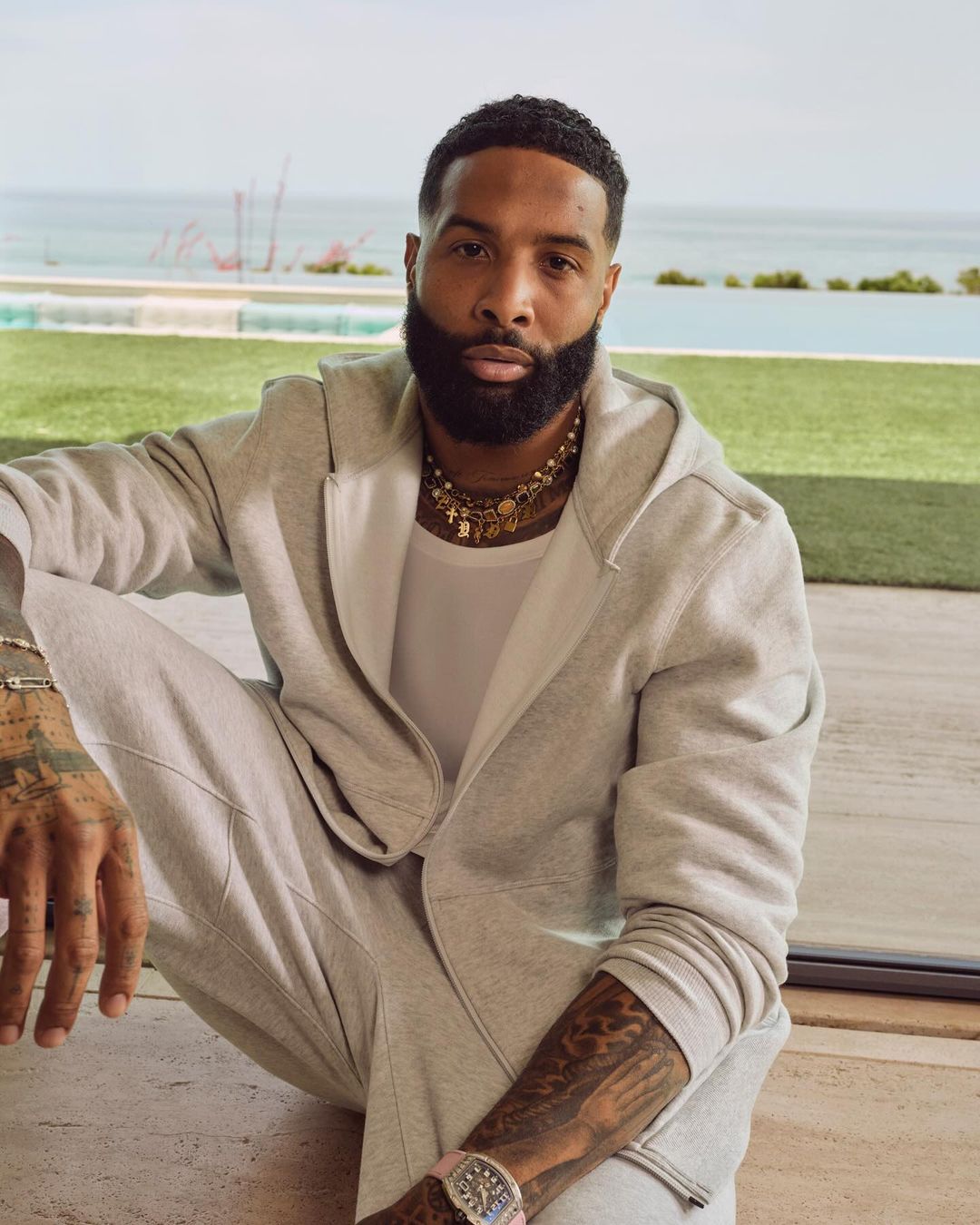 Adult male in grey sweatsuit posing outdoors with ocean and sky background. Man with beard and tattoos, wearing necklace and watch.