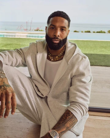 Adult male in grey sweatsuit posing outdoors with ocean and sky background. Man with beard and tattoos, wearing necklace and watch.