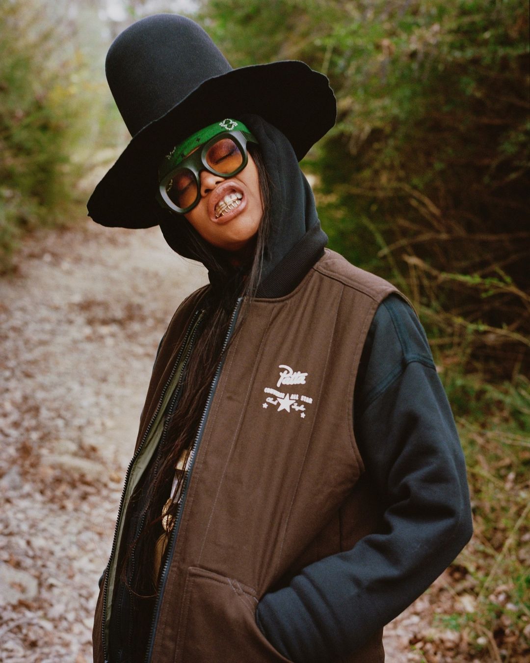 A person in a brown vest and black hat poses on a trail in a forest, wearing green sunglasses and a headscarf, smiling slightly.