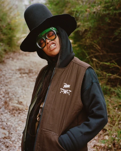 A person in a brown vest and black hat poses on a trail in a forest, wearing green sunglasses and a headscarf, smiling slightly.
