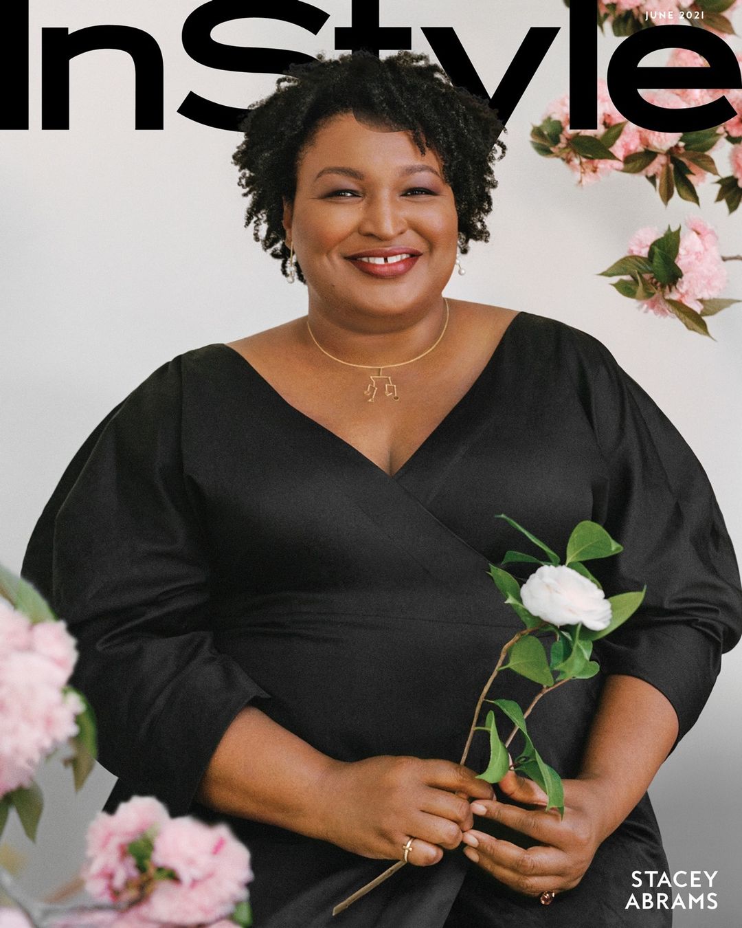 Stacey Abrams on magazine cover smiling, wearing a black dress and gold necklace, holding a white flower with pink floral background.