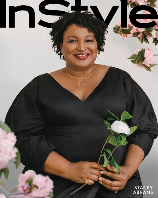 Stacey Abrams on magazine cover smiling, wearing a black dress and gold necklace, holding a white flower with pink floral background.
