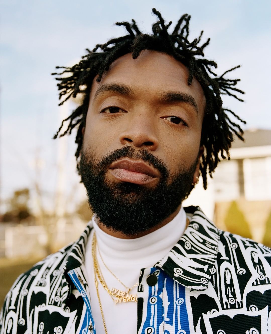 Close-up portrait of an adult Black man with dreadlocks, a beard, gold chains, and a patterned shirt set against an outdoor scene.