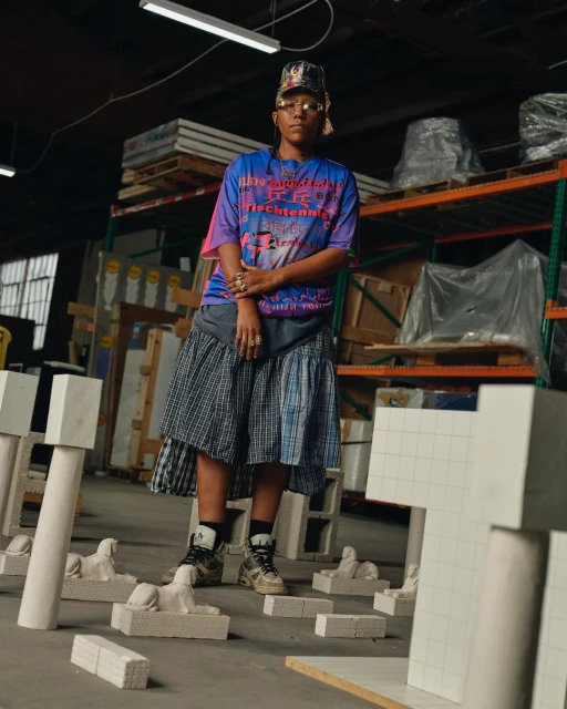 Full-length shot of an adult woman in a blue graphic tee, plaid skirt, and hat, posing indoors among white sculptural objects and warehouse shelving.