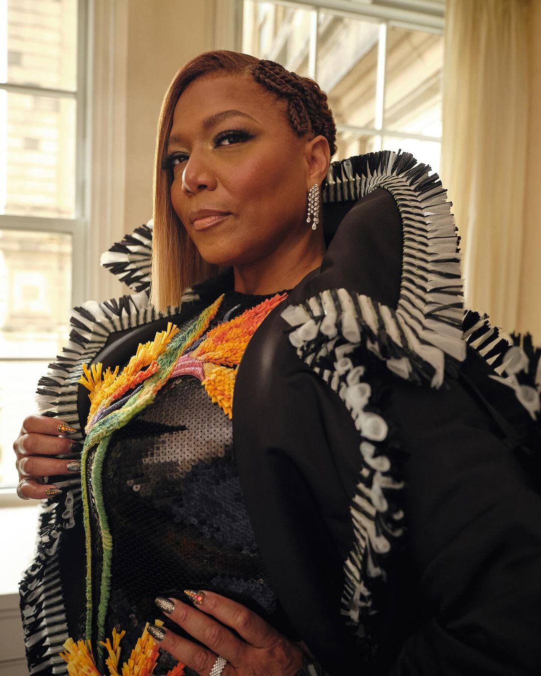 Headshot of Queen Latifah in a black dress with a vibrant floral design, accessorized with diamond earrings and rings, posing indoors.
