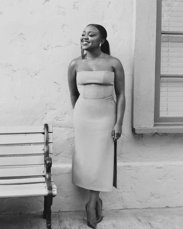 Black and white portrait of a smiling woman in a strapless midi dress standing outdoors near a wooden bench.