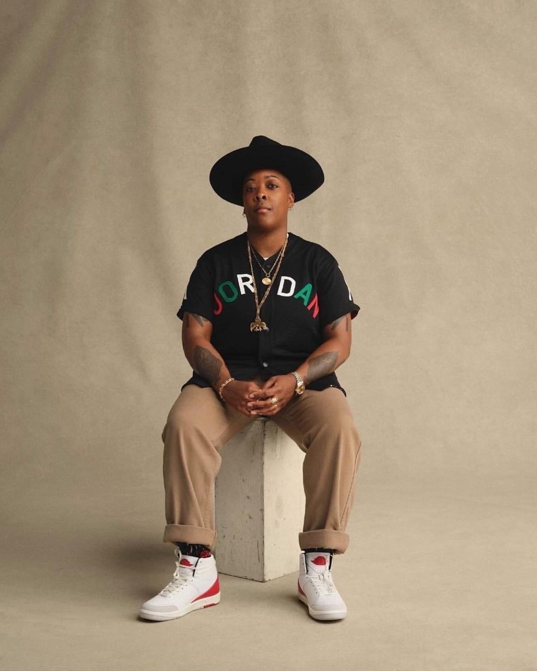 A person in a black hat and Jordan shirt is seated on a white block in a photo studio with a tan backdrop, wearing white sneakers.