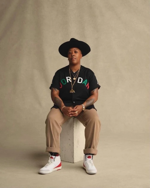 A person in a black hat and Jordan shirt is seated on a white block in a photo studio with a tan backdrop, wearing white sneakers.