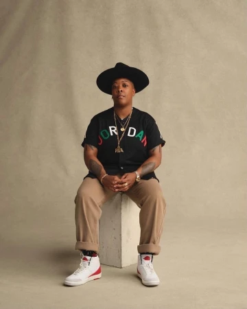 A person in a black hat and Jordan shirt is seated on a white block in a photo studio with a tan backdrop, wearing white sneakers.