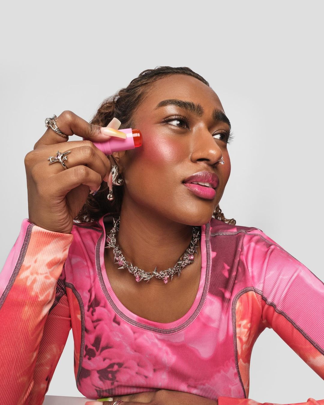 Close-up of an adult woman applying pink blush to her cheek with a lipstick-like applicator; she's wearing pink top and necklace.