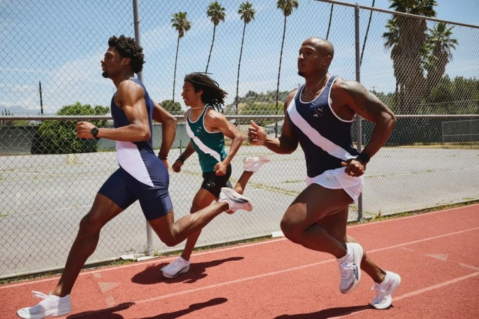 Three adult male athletes in running attire sprint on an outdoor track on a sunny day, palm trees and a chain-link fence in the background.