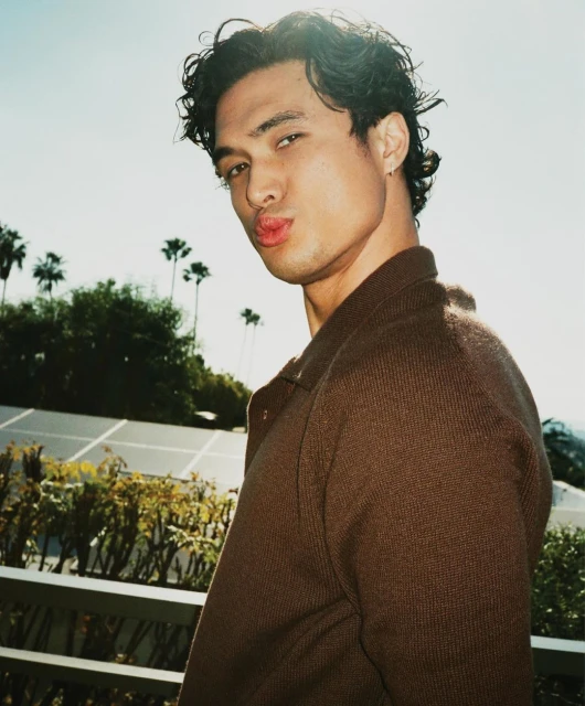 Portrait of a young adult male with curly dark hair, wearing a brown jacket, puckering his lips with palm trees and clear sky in background.