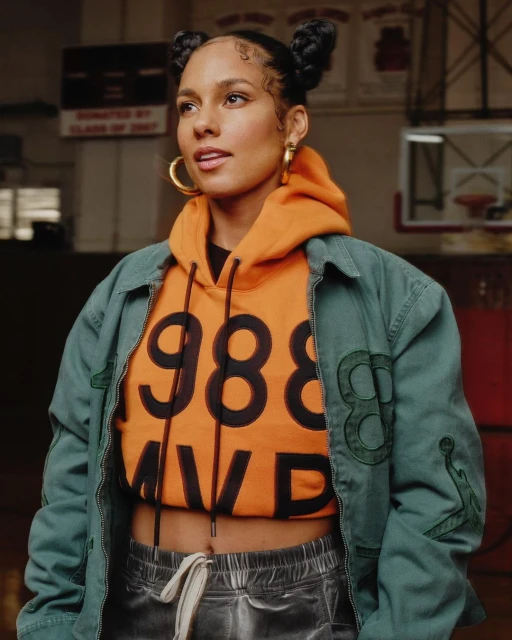 Alicia Keys poses in a teal jacket over an orange hoodie with '1988 MVP' text, against a gym wall with a basketball hoop.