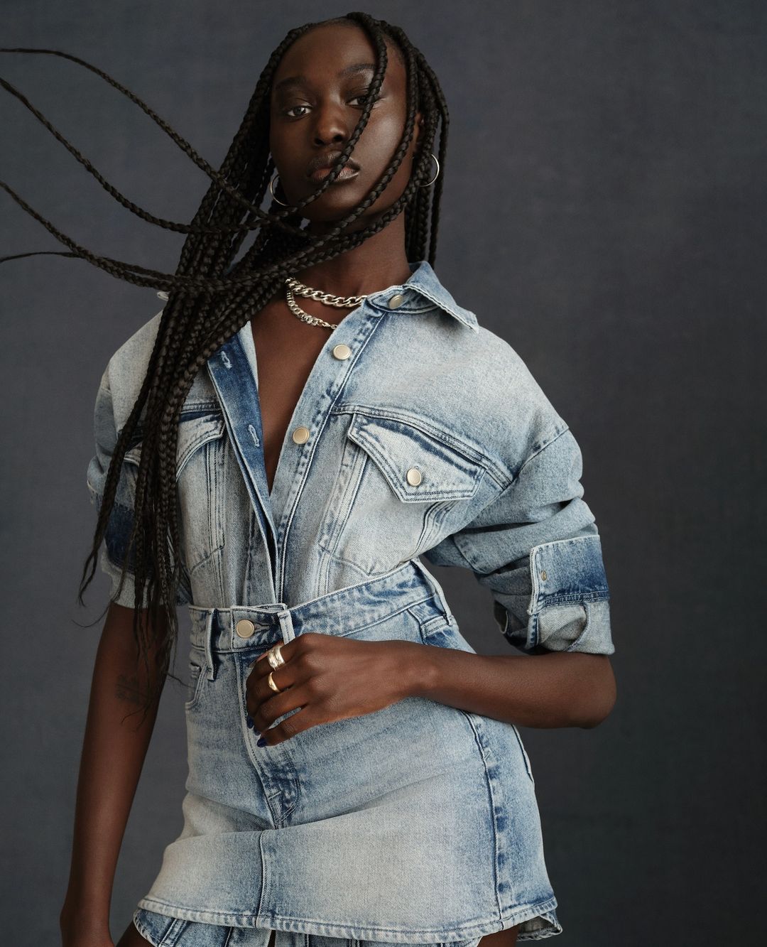 Portrait of a Black woman wearing a light blue denim jacket and skirt set, posing against a dark gray backdrop in a fashion editorial shot.