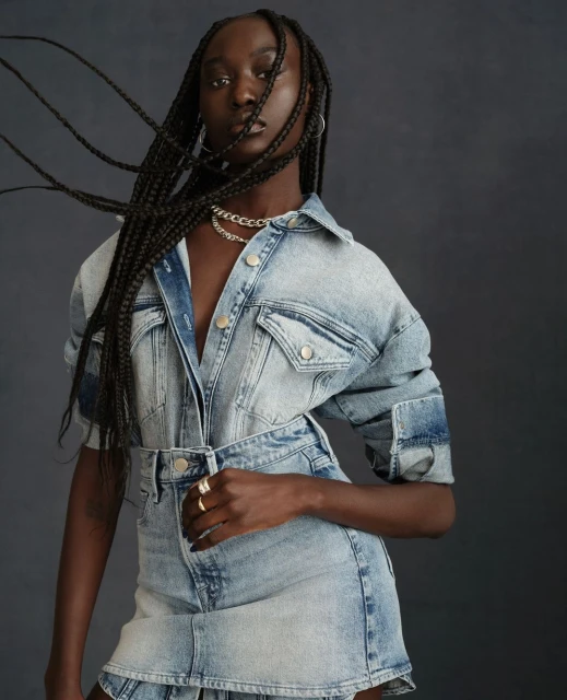 Portrait of a Black woman wearing a light blue denim jacket and skirt set, posing against a dark gray backdrop in a fashion editorial shot.