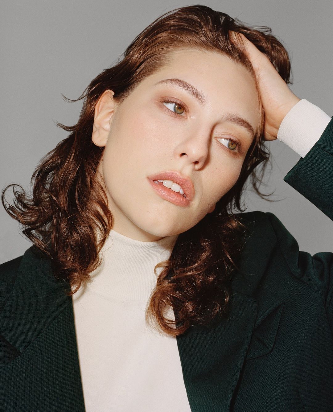 Portrait of a brunette adult woman with her hand in her hair, wearing a green blazer and a white turtleneck, looking off camera.