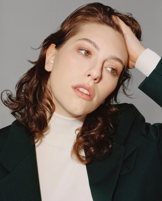 Portrait of a brunette adult woman with her hand in her hair, wearing a green blazer and a white turtleneck, looking off camera.