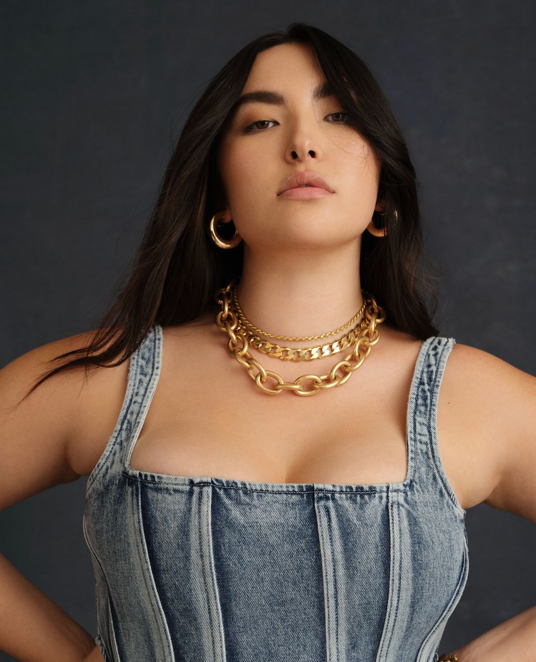 Portrait of a woman with dark hair wearing a denim top and gold jewellery against a neutral background in soft, diffused lighting.