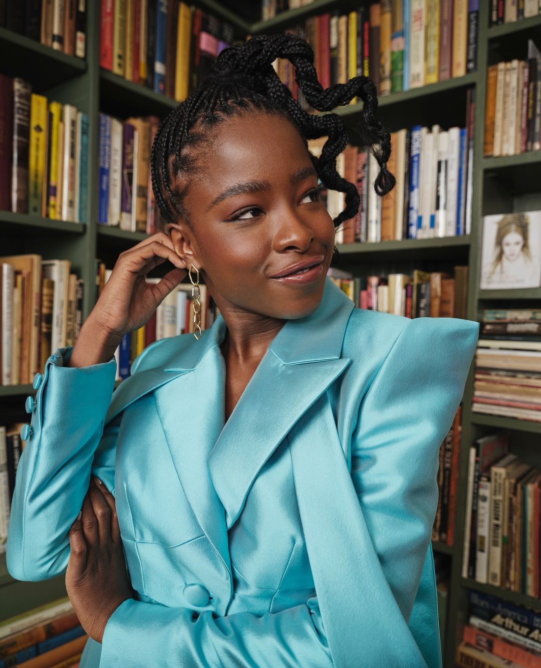 Portrait of a black woman wearing a bright turquoise blazer standing in front of a bookshelf filled with books.