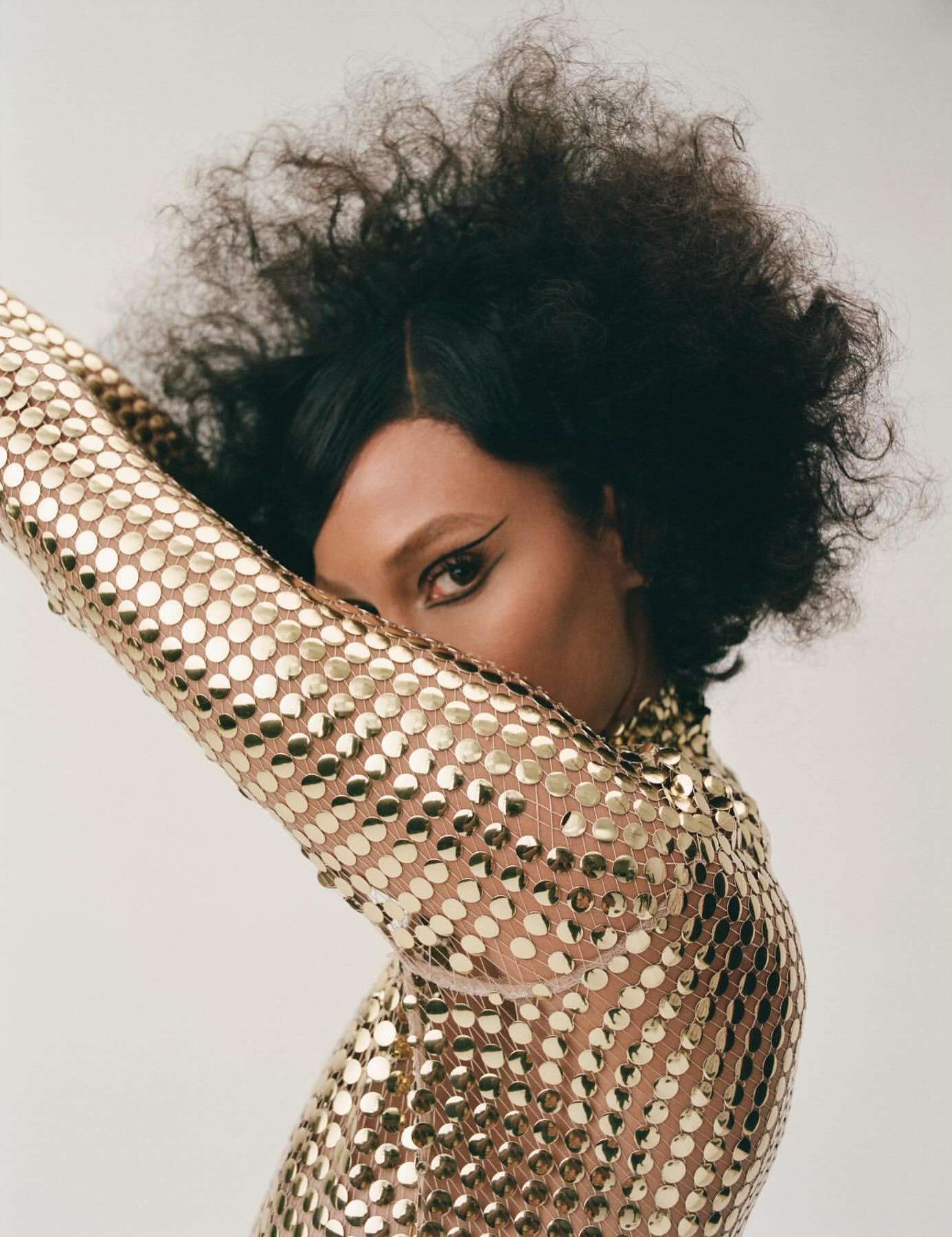 Adult woman with a voluminous afro hairstyle poses wearing a gold sequin dress. The photo is taken indoors against a neutral backdrop.