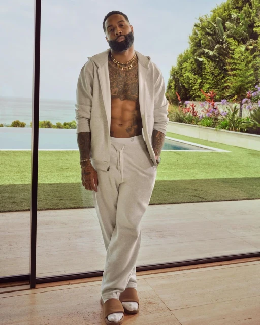 Athletic African-American male poses in a light-colored tracksuit and sandals against a modern residence with an ocean view, capturing a lifestyle scene.