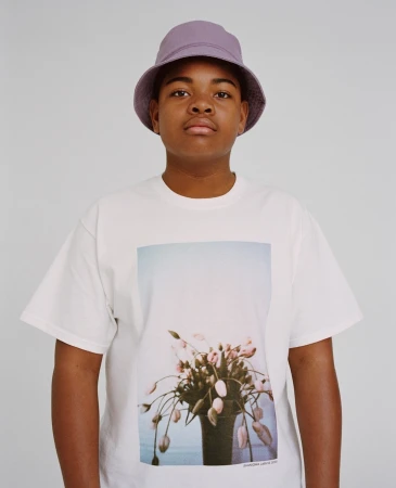Close-up of an African-American male wearing a lavender bucket hat and a white t-shirt with a floral design posing indoors.