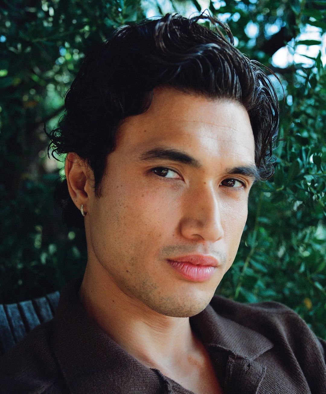 Close-up portrait of an Asian man with dark wavy hair wearing a brown collared shirt, set against a backdrop of green foliage.