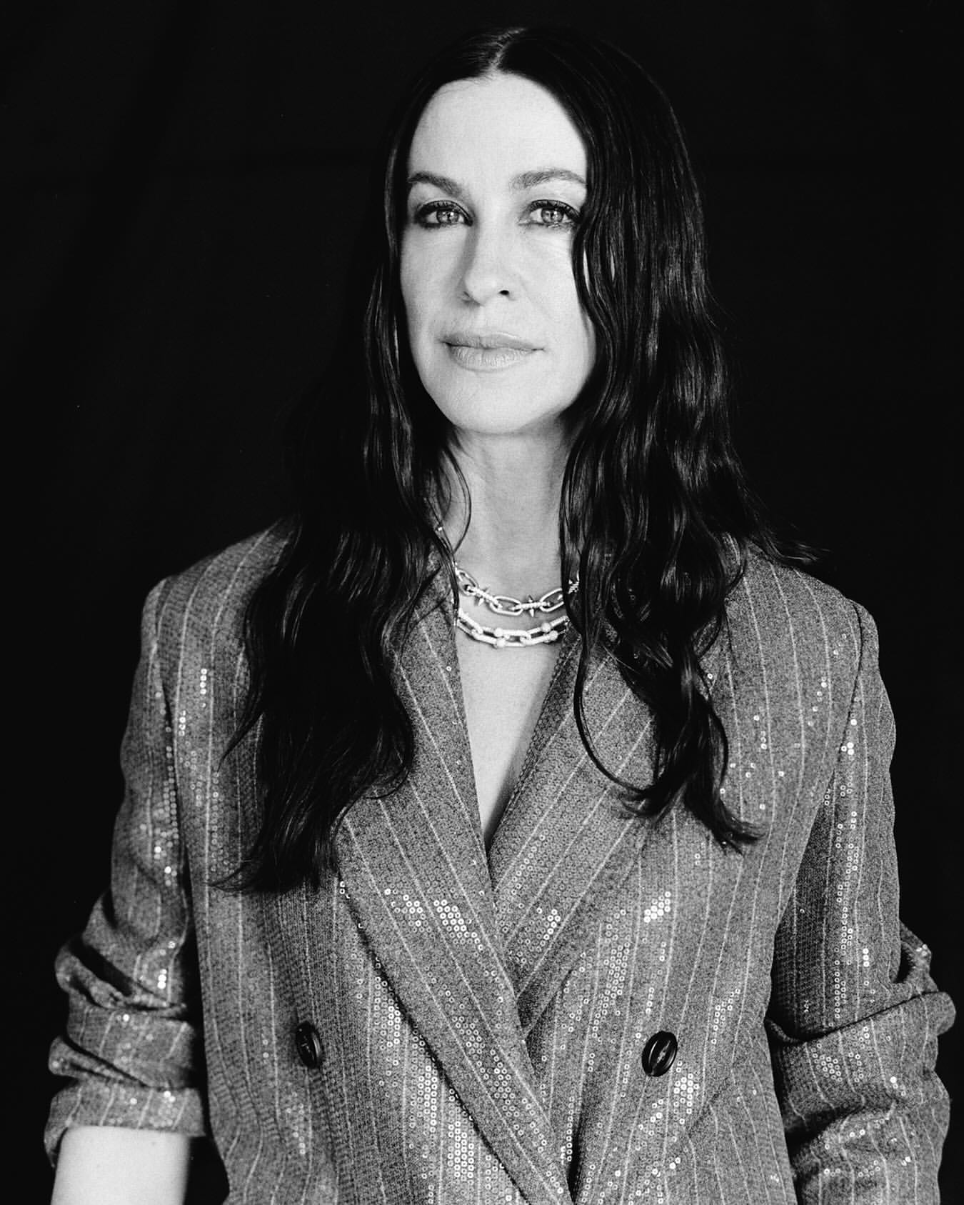 Monochrome portrait of a woman with long dark wavy hair wearing a sequined pinstripe blazer and layered chain necklace.