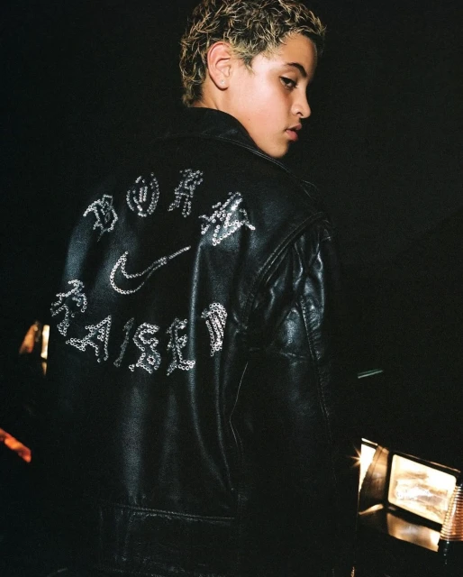 Teenager in a black leather jacket with embroidered brand logos and text, photographed in a low-key, indoor setting.