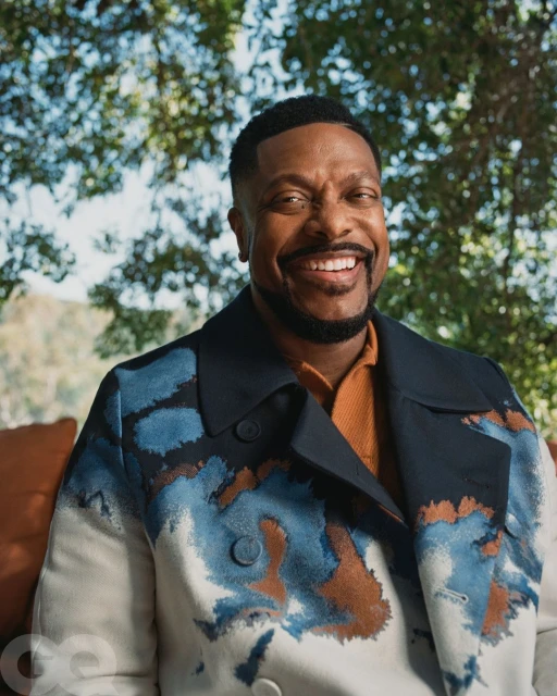 Portrait of actor Chris Tucker smiling in a patterned jacket outdoors with trees and foliage in the background in natural light.