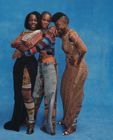 Three women embrace indoors against blue backdrop. They are wearing vibrant, patterned tops and denim, smiling, in a medium studio shot.