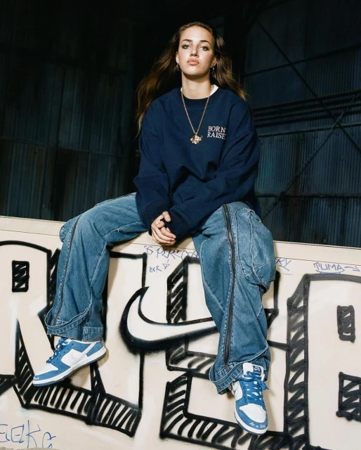 Young adult woman poses in blue jeans and sweatshirt, seated on graffiti-covered wall outdoors in an urban setting with blue shoes.