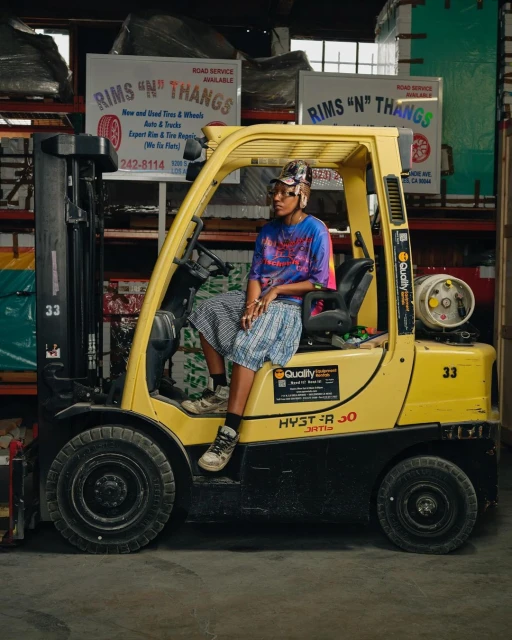 An adult female sits on a yellow forklift inside a tire warehouse with signage for auto services visible in the background. 