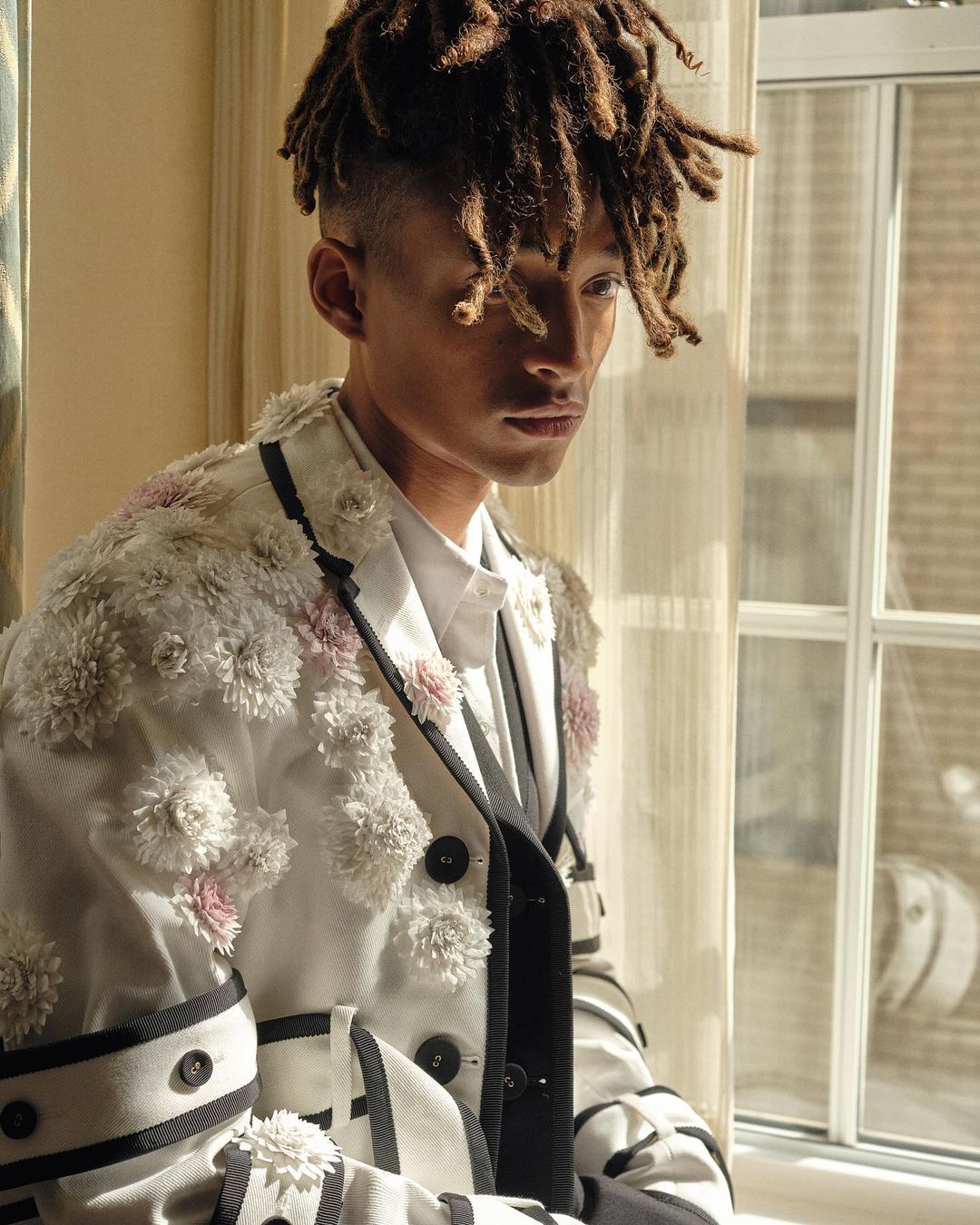 Portrait of a young adult with dreadlocks wearing a white jacket decorated with flowers, looking off to the side by a window.