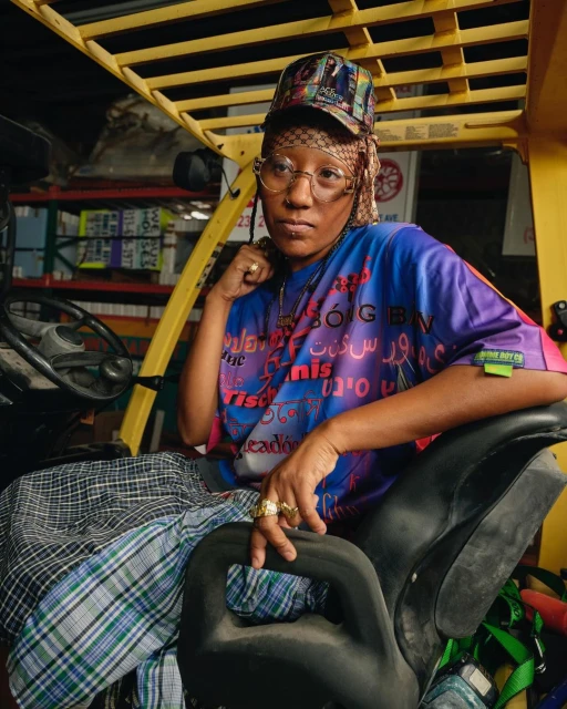 Person in colorful shirt and hat sitting on a forklift in an industrial setting with boxes and equipment visible in the background.