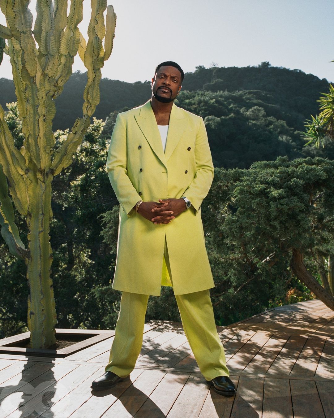 Portrait of an adult male in a chartreuse suit, standing outdoors with greenery and a cactus-like tree in the background.