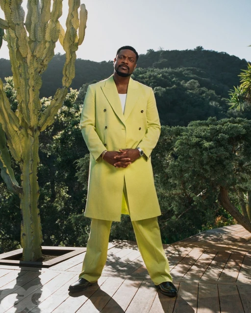 Portrait of an adult male in a chartreuse suit, standing outdoors with greenery and a cactus-like tree in the background.