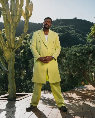 Portrait of an adult male in a chartreuse suit, standing outdoors with greenery and a cactus-like tree in the background.