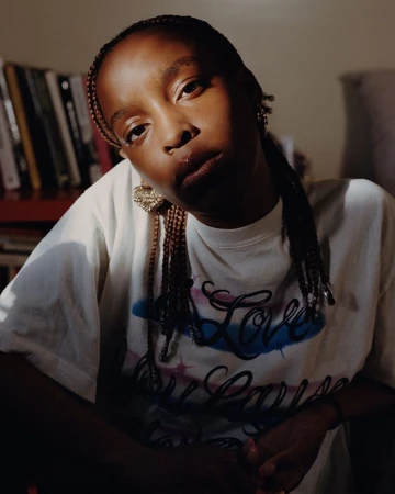 Portrait of a Black woman with braided hair wearing a white graphic t-shirt indoors with bookshelves visible in the background.