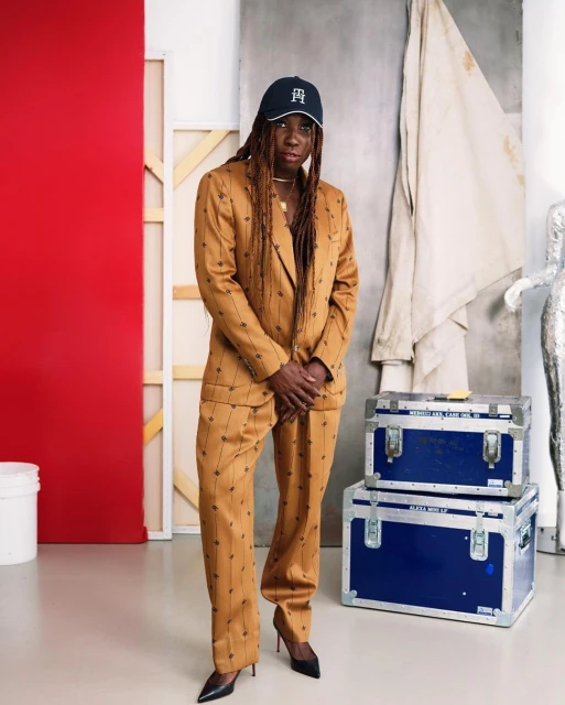 Stylish woman in a brown suit and cap poses in front of a painted backdrop inside an art studio, with metal boxes on the floor.