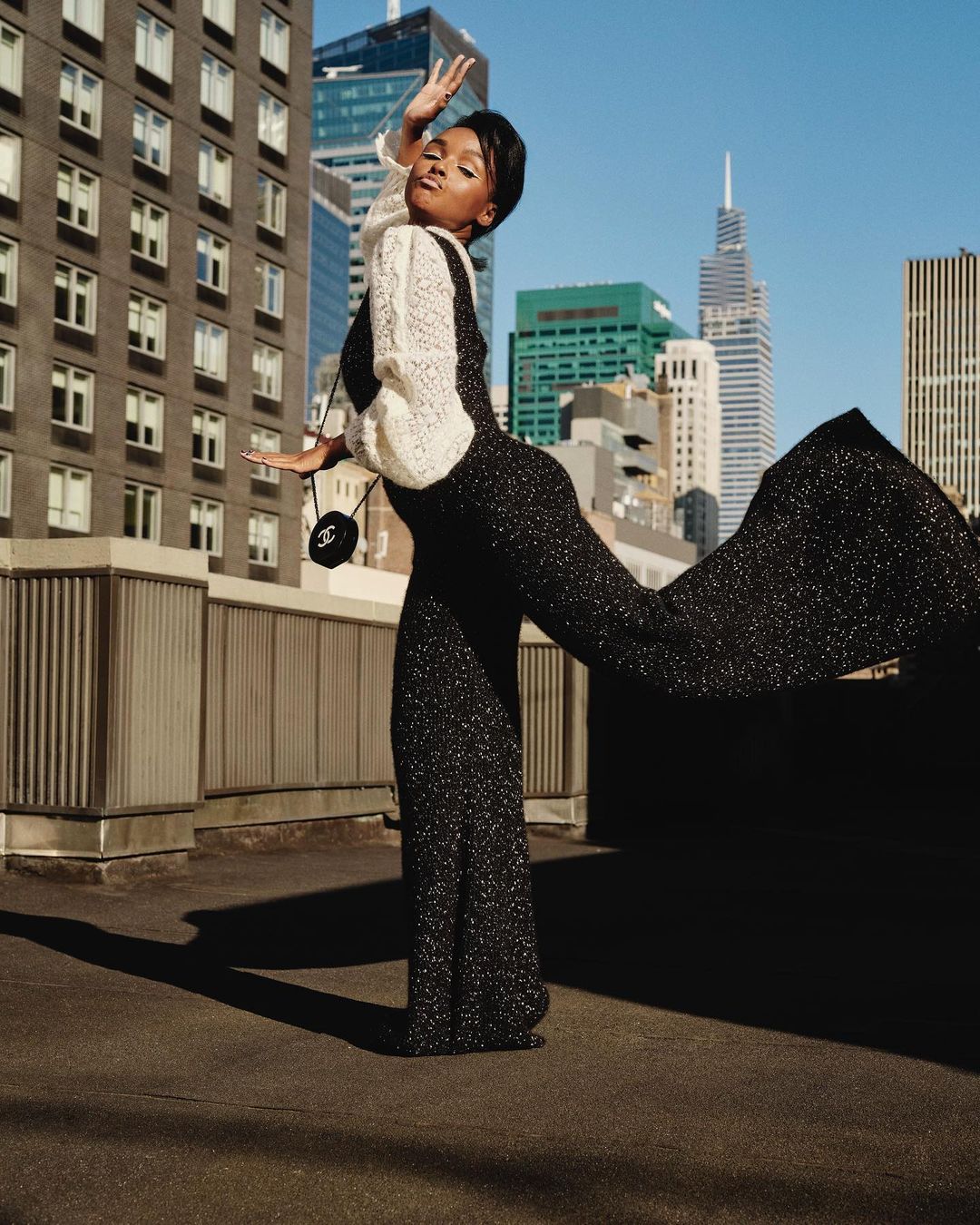 A fashionably dressed Black woman poses on a rooftop with city buildings in the background, wearing a stylish jumpsuit and purse.